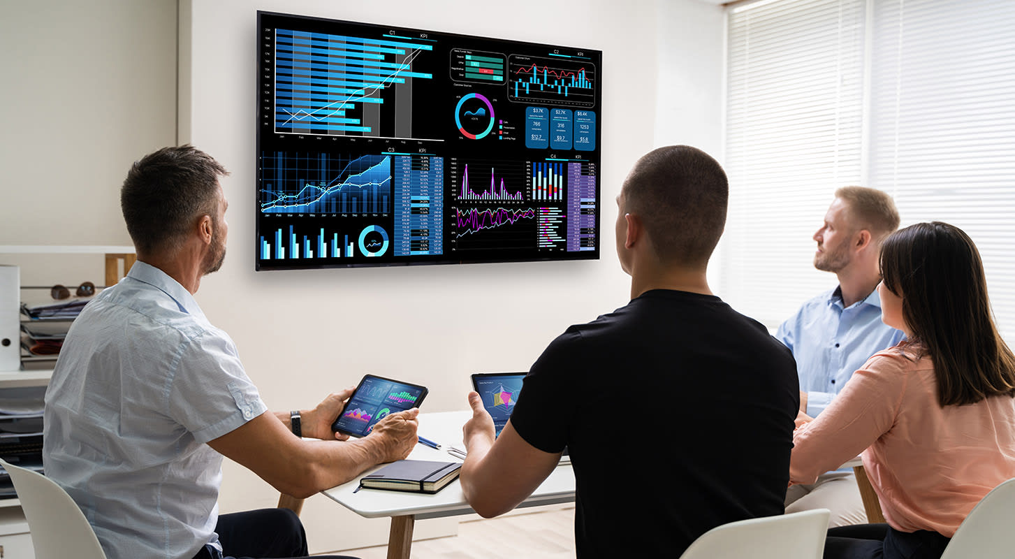 A diverse group of people collaborates around a table, discussing insights displayed on a large screen in a meeting setting.